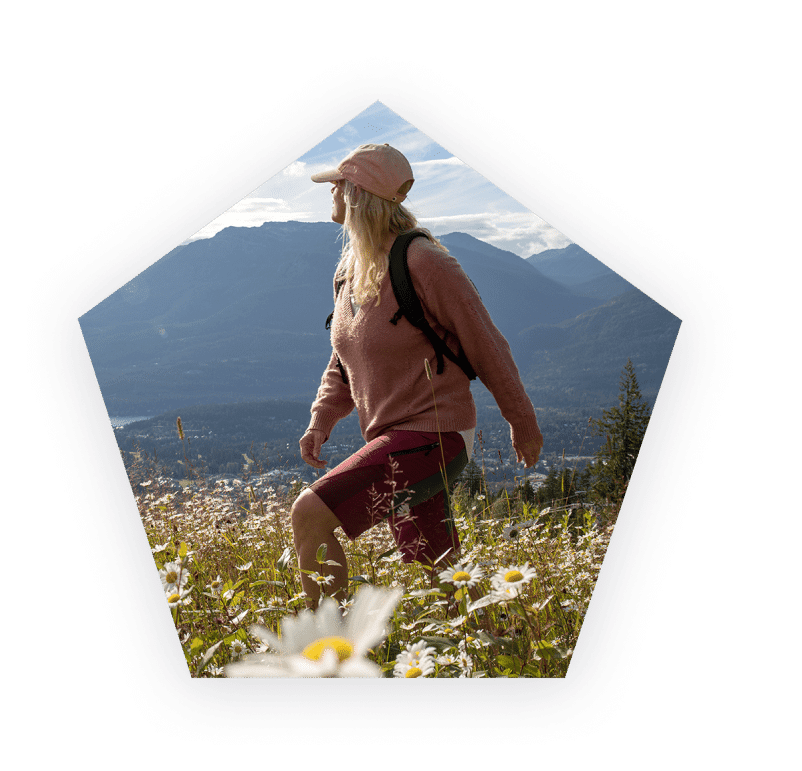 Woman wearing a hat hiking through a field of flowers and mountains in the background.